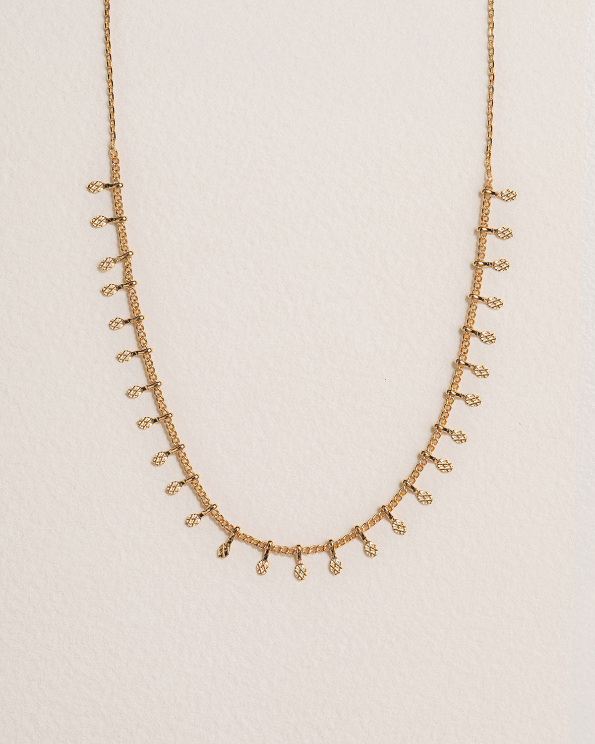 A 18k gold-plated necklace with vertical sequins on a fine chainayed against a beige background.